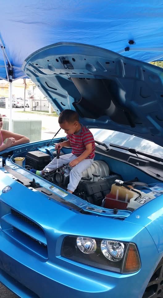 A kid sitting in the engine bay of a blue Dodge Charger, holding a screwdriver
